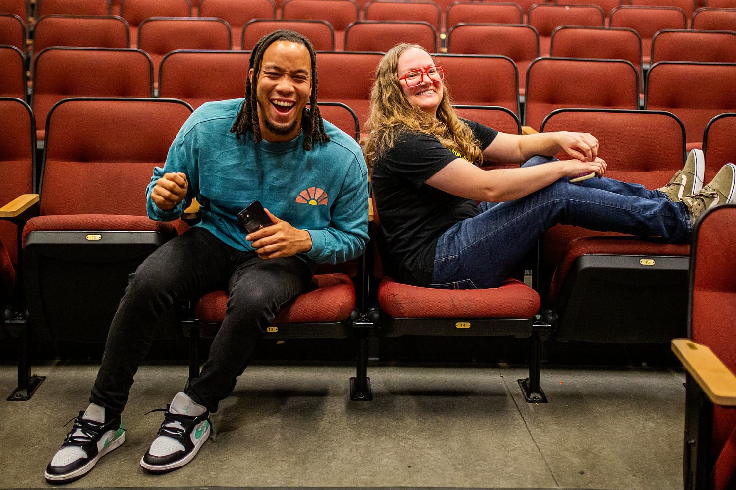 A man in a blue shirt and a women in a black shirt sitting and laughing in a row of red seats.