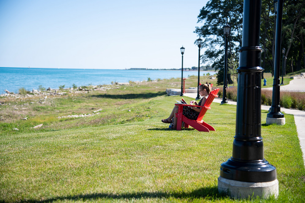 A student studies in a red Adirondack chair with a lake view.